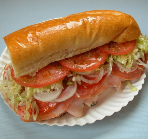 A large sandwich on a white plate, filled with layers of lettuce, tomatoes, onions, and sliced meats in a soft bun.