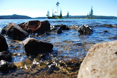A serene lake scene with clear water, rocky shoreline, and distant trees under a bright blue sky.