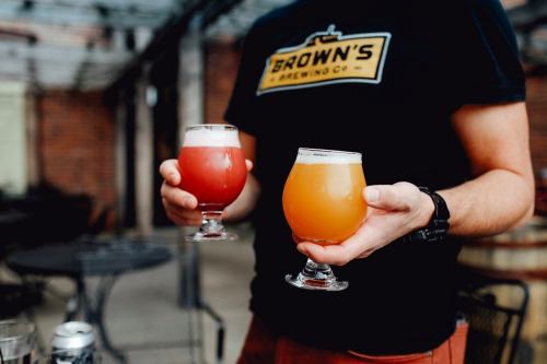 A person holds two craft beer glasses, one with a red drink and the other with a golden drink, in a brewery setting.