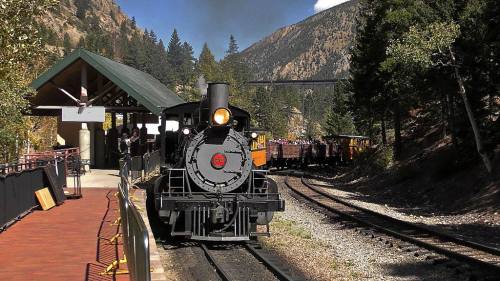 A vintage steam train approaches a station surrounded by mountains and trees, with a train track curving in the background.