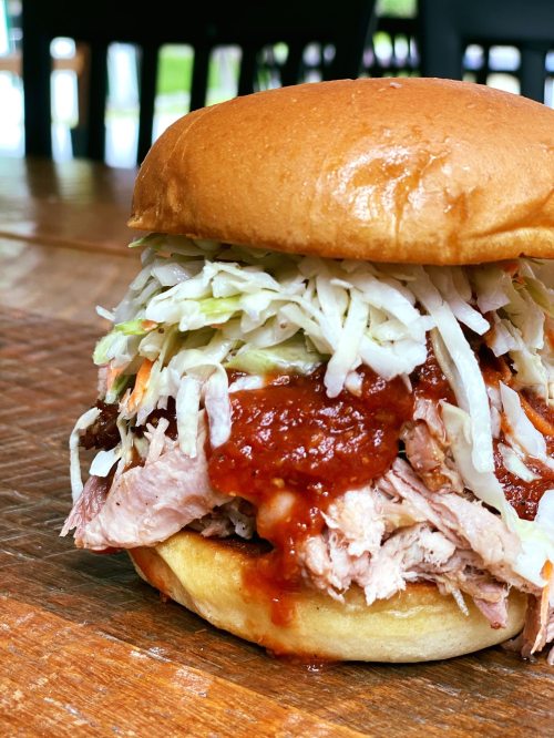 A close-up of a pulled pork sandwich topped with coleslaw and barbecue sauce, served on a soft bun.
