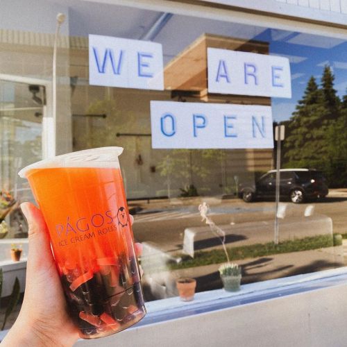 A hand holding a drink in front of a shop window that says "WE ARE OPEN." The drink has colorful layers and toppings.