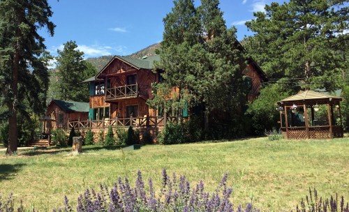 A rustic wooden lodge surrounded by trees and a gazebo, set against a backdrop of mountains and blue sky.