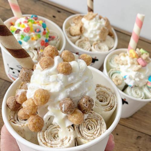 A hand holding a cup of rolled ice cream topped with whipped cream and cereal, surrounded by other dessert cups.