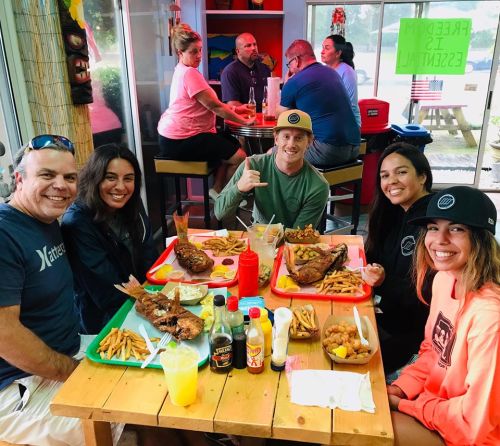 A group of five friends enjoying a meal with fried fish, fries, and drinks at a lively restaurant.