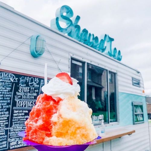 A colorful snow cone topped with whipped cream and a strawberry, in front of a food truck named "Shaved Ice."