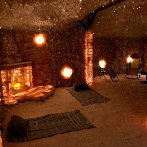 A cozy, dimly lit salt room with stone walls, a fireplace, and reclining chairs on sandy floors.