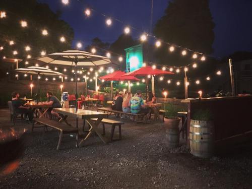 Outdoor dining area at night, featuring string lights, umbrellas, and people enjoying food and drinks at wooden tables.