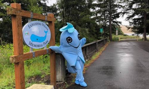 A blue whale mascot leans against a sign for Exploding Whales Memorial Park, surrounded by trees and a rainy path.
