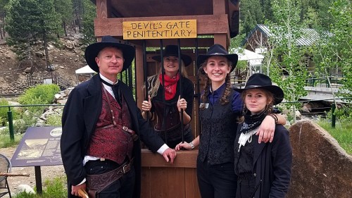 Four people in Western attire pose at the Devil's Gate Penitentiary sign, surrounded by trees and a rustic setting.