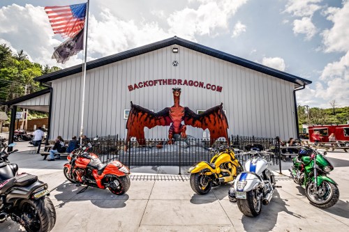 A building with a dragon mural, surrounded by colorful motorcycles and flags, under a bright blue sky.