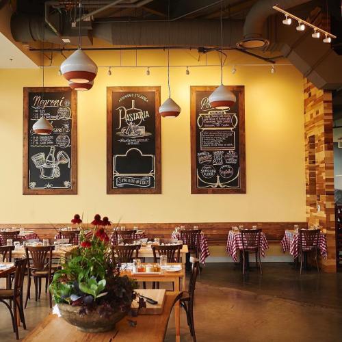 Interior of a restaurant featuring wooden tables, checkered tablecloths, and chalkboard menus on yellow walls.