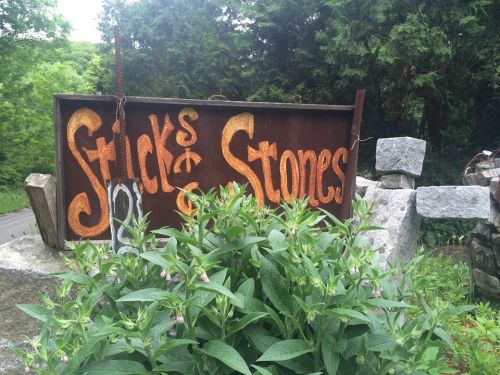 Rusty sign reading "Sticks & Stones" surrounded by greenery and stone structures.