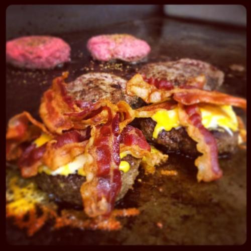 Two juicy burgers topped with melted cheese and crispy bacon, sizzling on a grill, with pink patties in the background.