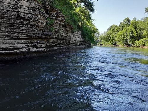 A serene river flows past rocky cliffs and lush greenery under a clear blue sky.