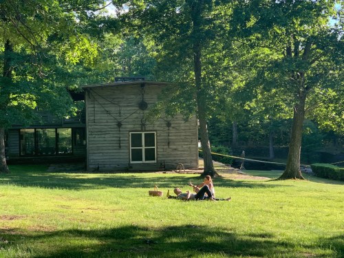 A person sits on the grass in front of a wooden building, surrounded by trees and a peaceful outdoor setting.