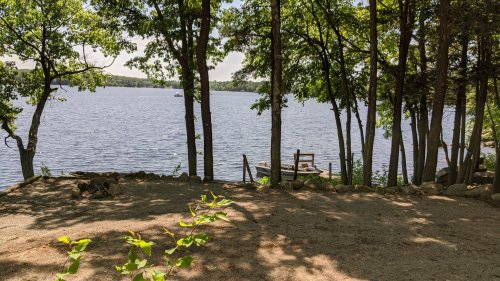 A serene lakeside view framed by trees, with a small dock and boats on the calm water under a clear sky.