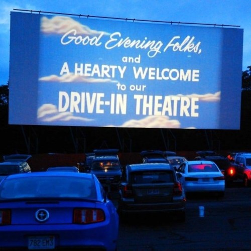 A drive-in theater screen displays a welcome message as cars are parked in front under a twilight sky.