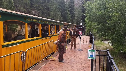 A vintage train at a station with passengers, surrounded by trees and a sign reminding to wear masks.
