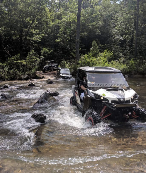 Two off-road vehicles navigate through a rocky stream surrounded by lush greenery.