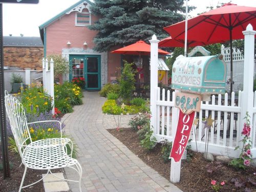 Charming cookie shop with a colorful garden, white picket fence, and red umbrellas over outdoor seating. Sign reads "OPEN."