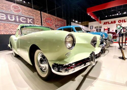 A vintage light green car displayed in a retro-themed museum with brick walls and classic signage in the background.