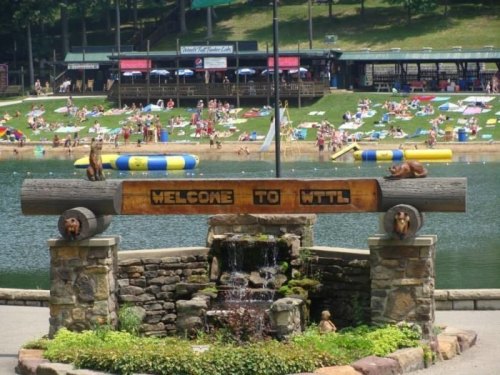 A wooden sign reading "Welcome to WTTL" with a waterfall, surrounded by people enjoying a lake and beach area.