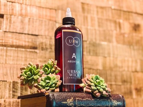 A bottle of Urban Wick Absinthe fragrance sits on a textured surface, surrounded by small succulent plants.