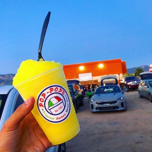 A hand holds a bright yellow snow cone in a cup, with cars and a building in the background during twilight.