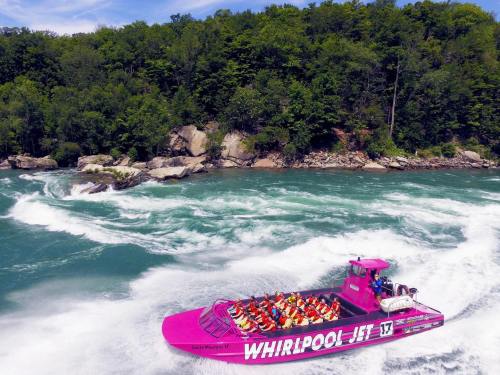 A bright pink jet boat with passengers navigates through swirling waters near a rocky shoreline and lush greenery.