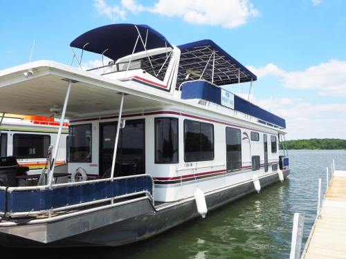 A large houseboat docked at a marina, featuring a spacious upper deck and surrounded by calm water under a blue sky.