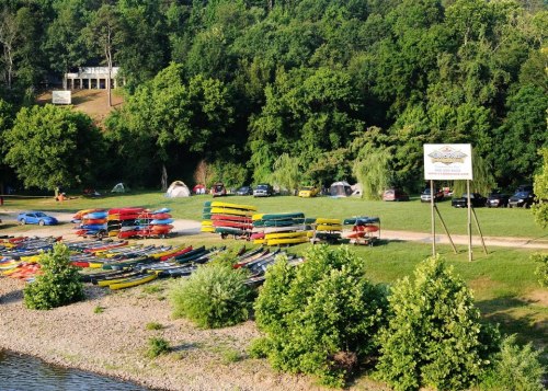 A riverside scene with colorful kayaks, tents, and cars surrounded by lush green trees.