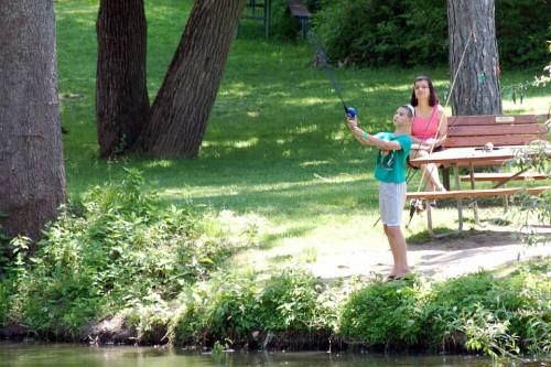 A child fishing by a pond while an adult sits on a bench nearby, surrounded by greenery.