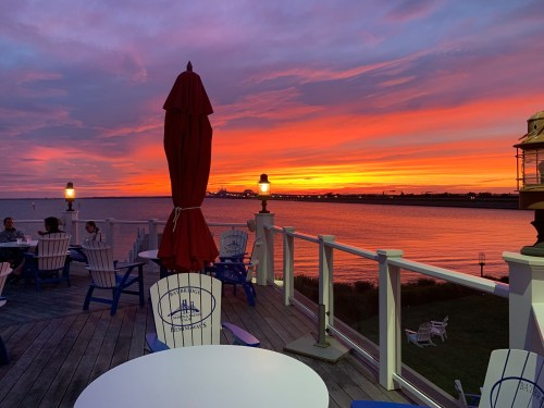 A vibrant sunset over a calm waterfront, with outdoor seating and umbrellas in the foreground.