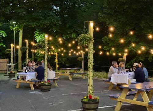Outdoor dining area with string lights, featuring tables set for dinner and guests enjoying a meal in a lush setting.