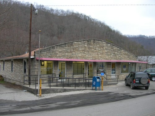 A stone building with a pink awning, serving as a post office, surrounded by a rural landscape and mountains.