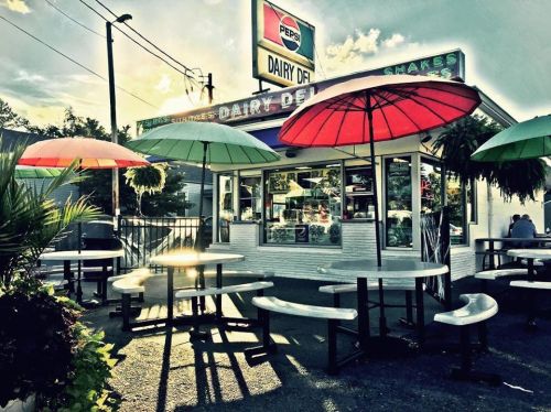 A colorful dairy stand with umbrellas over outdoor seating, surrounded by greenery and a bright sky.