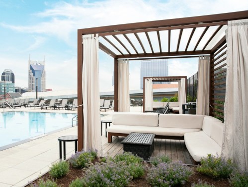 Rooftop pool area with lounge seating and city skyline in the background, featuring modern cabanas and greenery.