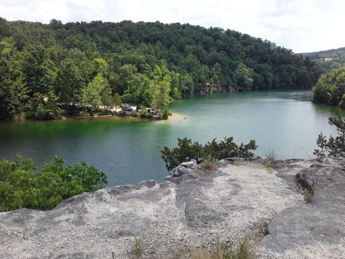 A serene lake surrounded by lush green trees and rocky cliffs under a partly cloudy sky.