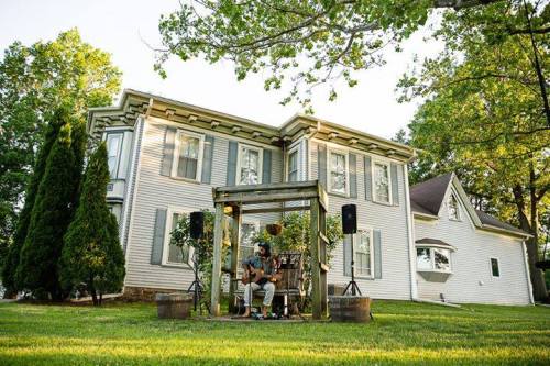 A musician performs under a wooden pergola in front of a large, white house surrounded by trees.