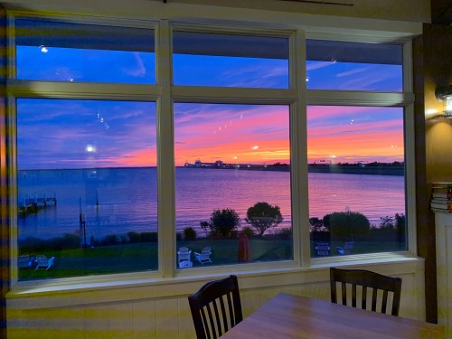 A vibrant sunset over a calm waterfront, viewed through a large window with wooden chairs in the foreground.