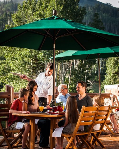 A chef serves food to a family at an outdoor dining table under green umbrellas, surrounded by trees and mountains.