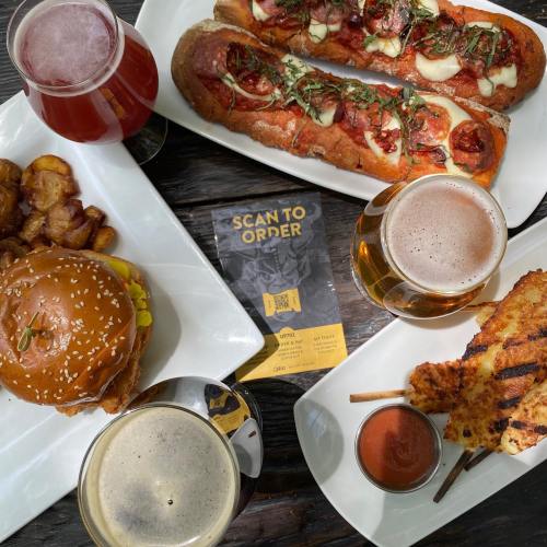 A spread of food and drinks including burgers, hot dogs, skewers, and craft beers on a wooden table.