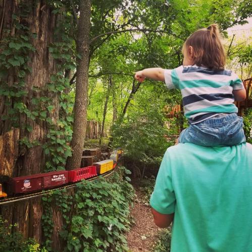 A child on an adult's shoulders points at a miniature train running through a lush, green forested area.