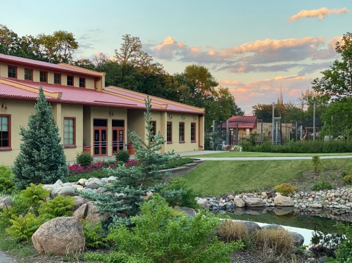 A landscaped area featuring a building with a red roof, surrounded by greenery, rocks, and a pond under a colorful sky.