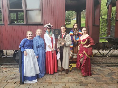 A group of six people in historical costumes poses in front of a vintage train car.