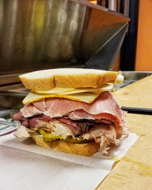 A towering sandwich with layers of various meats, cheese, and pickles, served between two slices of bread.
