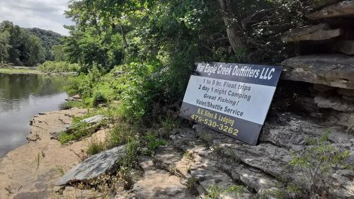 Sign for Eagle Creek Outfitters LLC near a river, promoting float trips, camping, and fishing services.
