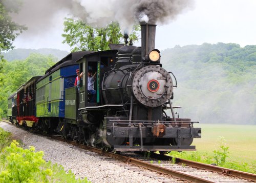 A vintage steam locomotive chugs along a scenic track, billowing smoke against a backdrop of green hills.