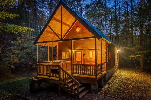 A cozy wooden cabin illuminated at dusk, surrounded by trees and a gravel path leading to the entrance.
